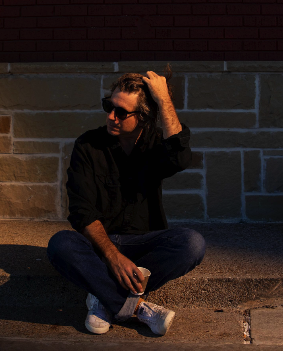 Jeffrey paradise sits on a curb with sunglasses looking away from the camera