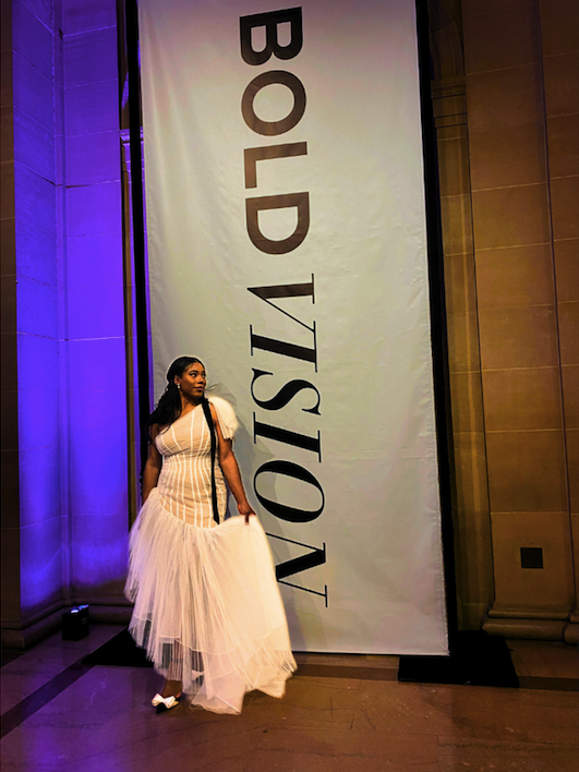 a shot of a Black woman in a long white gown with tulle bottom in front of a sign reading "bold vision"