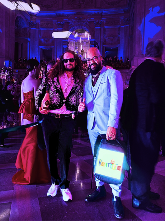 two men pose wearing flamboyantly adorned and colored suit ensembles