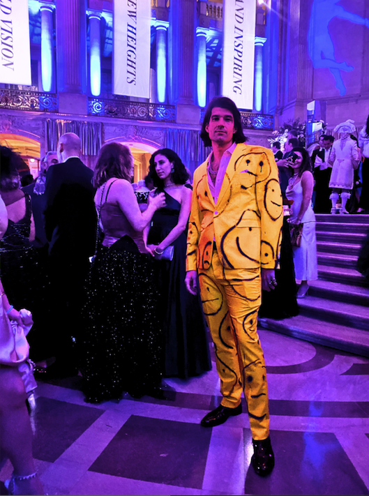 a man poses in front of the grand staircase in a yellow suit adorned with black smiley faces