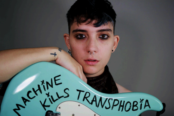 headshot of the artist holding a guitar with "this machine kills transphobia" written on it in black marker