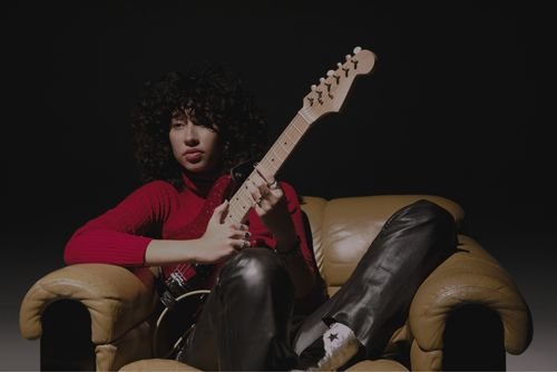 Towa sits with her guitar on a beige old fashion style leather love seat wearing a red top and brown leather pants