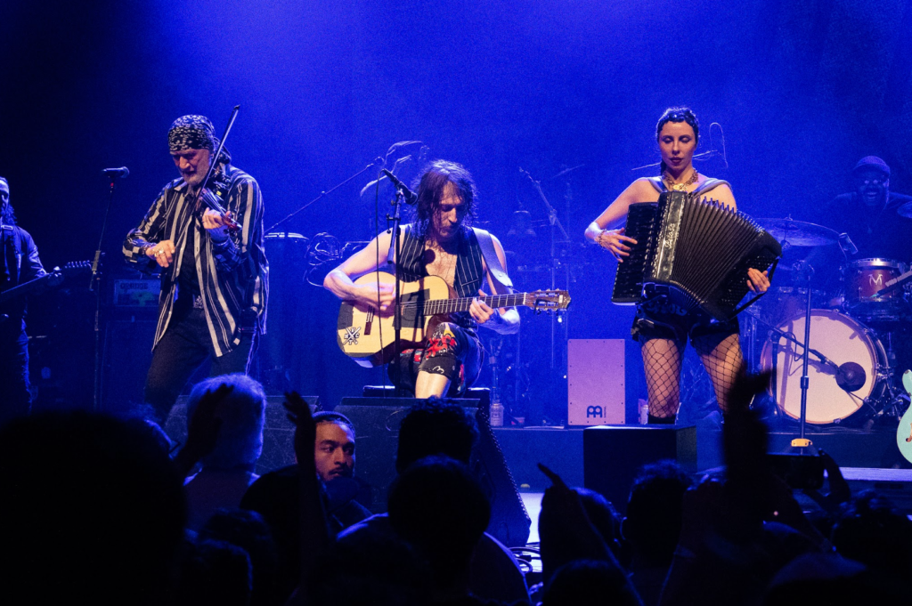 Gogol Bordello performing with acoustic guitar, violin, and accordion under blue lighting