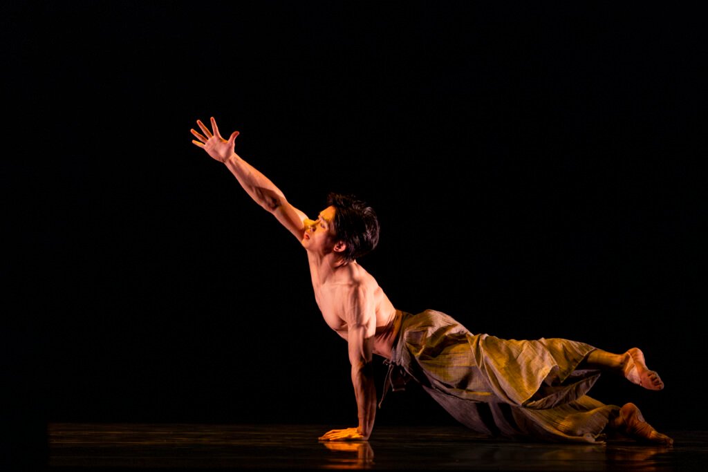 Male ballet dancer extends one arm upward while balancing low to the ground in a contemporary solo performance