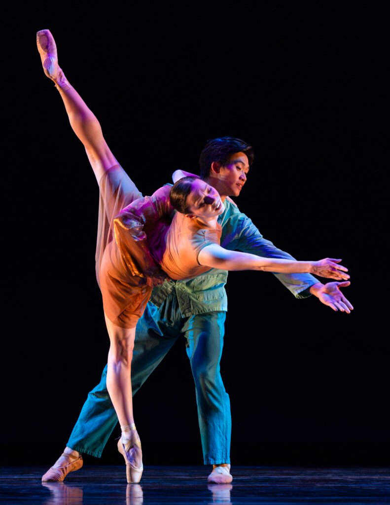 Ballet dancers Jazmine Quezada and Lawrence Chen perform a contemporary duet with extended leg line and supported reach on a dark stage