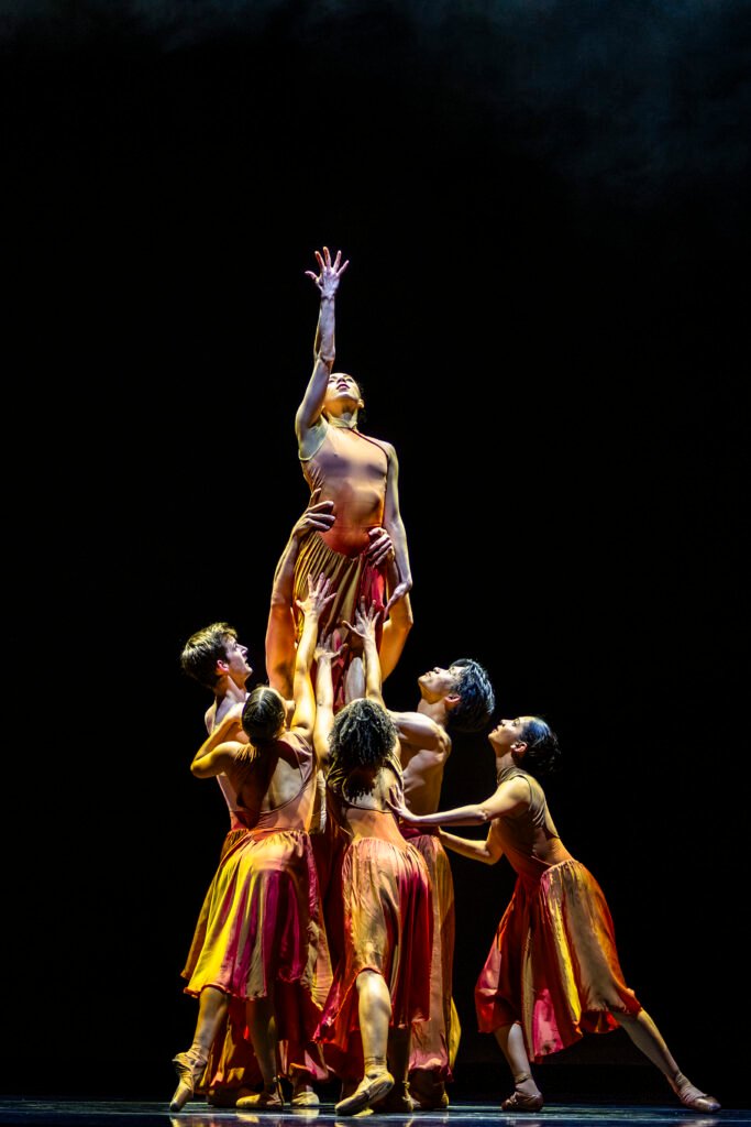 Group of ballet dancers lift a central performer reaching upward in a dramatic ensemble formation on a dark stage