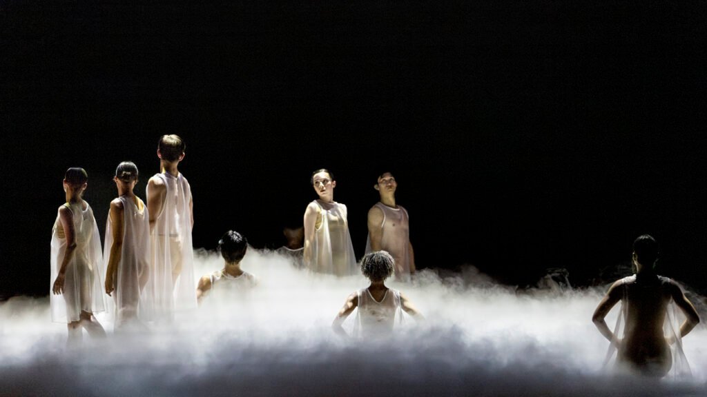 Ballet dancers stand in a low fog wearing light costumes, partially obscured in a dimly lit stage environment