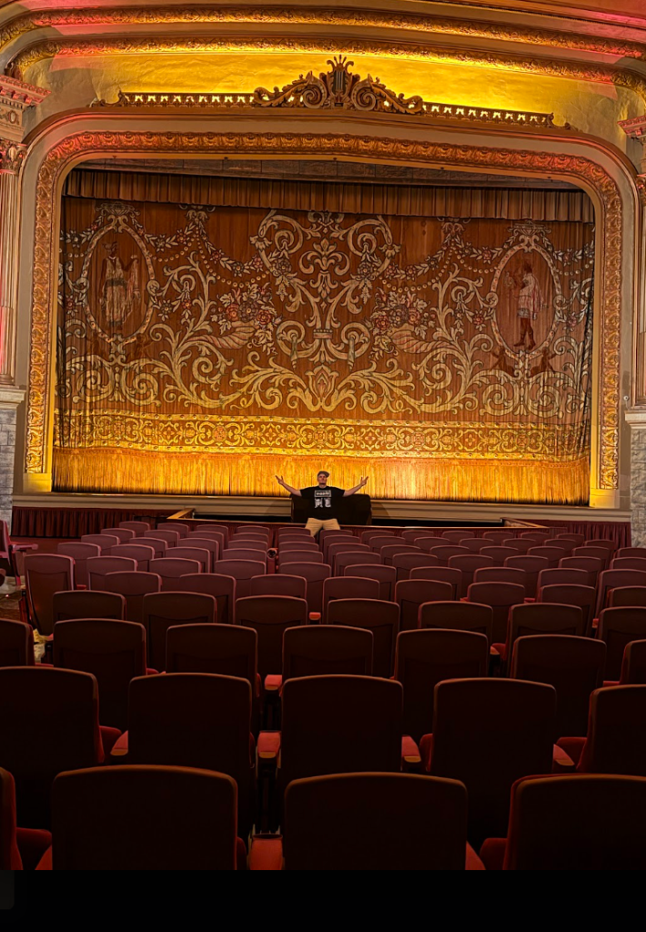 Skweezy Jibbs standing in front of the ornate stage curtain at the Grand Lake Theater in Oakland